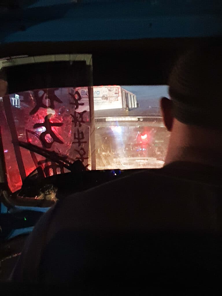 A male bajaji driver drives through Dar es Salaam's traffic at night. His windshield is broken and fixed with some huge sticker, entailing Chinese characters, a symbol of China in Tanzania.